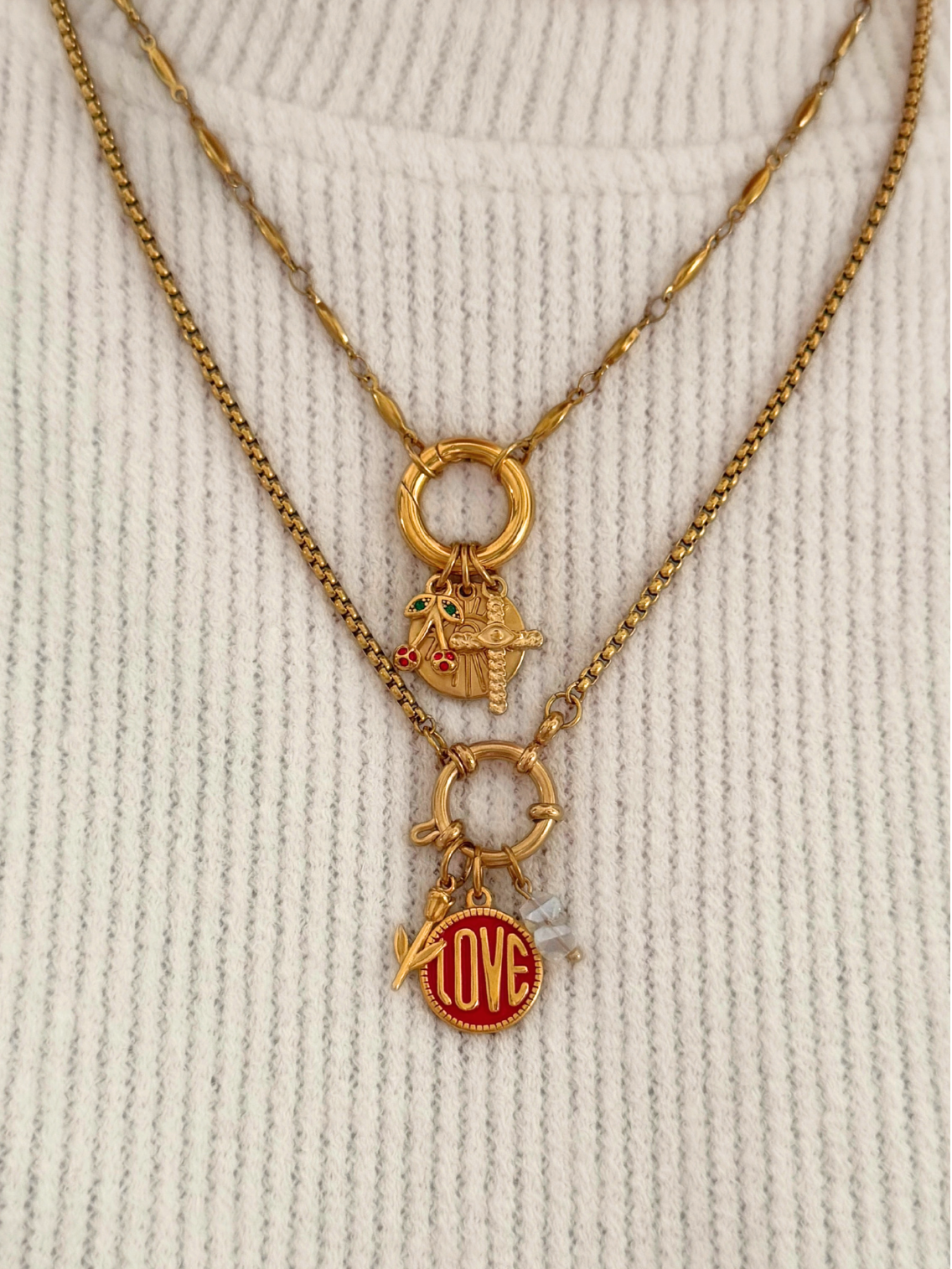 A close-up of a person wearing a layered gold necklace set over a cream ribbed knit sweater. The necklace features a top chain with a ring clasp and small charms, and a longer chain with a ring clasp, a tiny charm, and a red circular charm with gold lettering.